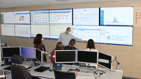 Besuch im Service Operation Centre (SOC): Die vier Schülerinnen stehen gemeinsam mit einem HZD-Mitarbeiter vor einer großen Wand mit zahlreichen Monitoren, dem sogenannten Cockpit. Der Kollege erläutert, was die verschiedenen Anzeigen bedeuten.