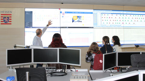 Besuch im Service Operation Centre (SOC) - Teil 2: Die vier Schülerinnen stehen gemeinsam mit einem HZD-Mitarbeiter vor der linken Seite des sogenannten Cockpits. Der Kollege deutet auf einen Monitor links oben und erläutert die Details